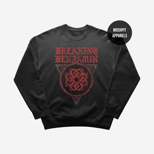 Può includere: Una felpa nera a girocollo con una grafica rossa dall'aspetto vissuto sul petto. Il testo recita BREAKING BENJAMIN sopra un disegno a nodo circolare racchiuso in un triangolo. Un badge circolare nero nell'angolo in alto a destra mostra MOSHPIT APPARELS.