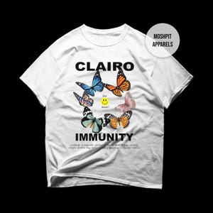 May include: A white short-sleeve t-shirt featuring the text CLAIRO IMMUNITY in black. The graphic includes five colorful butterflies arranged in a circle around a yellow smiley face with the text live music! and a tracklist at the bottom.