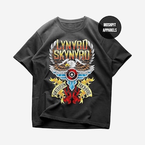 Pode incluir: Uma t-shirt preta de manga curta com uma impressão gráfica de uma águia-de-cabeça-branca com asas abertas, um escudo circular e o texto Lynyrd Skynyrd Southern Rock Roll. Um pequeno círculo preto contém o texto MOSHPIT APPARELS.