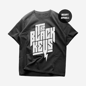 Può includere: Una t-shirt grigio antracite a maniche corte con una stampa grafica bianca dall'effetto consumato che recita THE BLACK KEYS con un fulmine. Un logo circolare nero nell'angolo in alto a destra contiene il testo bianco MOSHPIT APPARELS.