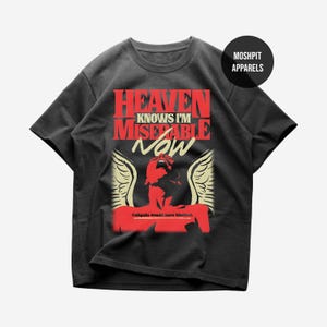 Puede incluir: Una camiseta negra de manga corta con un estampado gráfico en rojo y crema. El texto dice HEAVEN KNOWS I'M MISERABLE NOW, con la frase Caligula would have blushed bajo una silueta roja de una figura con alas. Se ve un logo de MOSHPIT APPARELS.