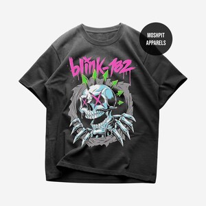 May include: A black short-sleeve t-shirt featuring a graphic of a skull with pink X-shaped eyes and green arrows emerging from its head. The text blink-182 is printed in pink above the skull, and a black circle with MOSHPIT APPARELS is visible.