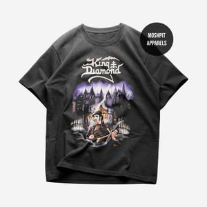 May include: A black short-sleeve t-shirt featuring a graphic print of King Diamond in front of a gothic castle gate. The design includes the text King Diamond in white, a figure holding a bone, and a purple sky. A black circle with MOSHPIT APPARELS is visible.