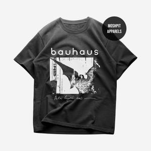 May include: A black short-sleeved t-shirt featuring a white graphic print of a bat-like figure. The text "bauhaus" appears above the graphic, and "Bela Lugosi's dead" is written below. A circular logo in the top right corner reads "MOSH PIT APPARELS".