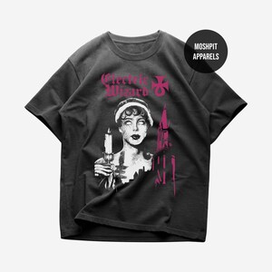 May include: A charcoal grey short-sleeve t-shirt featuring a graphic print in pink and white. The design includes the text Electric Wizard, a person holding a candle, and a building silhouette. A circular logo reads MOSHPIT APPARELS.