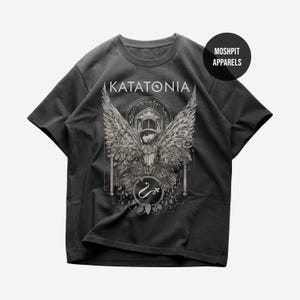 May include: A charcoal gray short-sleeve t-shirt featuring a graphic print with the text KATATONIA at the top. The design displays a large winged creature, a circular emblem with a symbol, and architectural elements in a light gray ink.