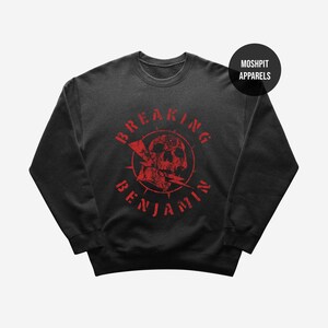 Può includere: Una felpa nera a maniche lunghe con una stampa grafica rossa raffigurante un teschio attraversato da un fulmine. Il testo "BREAKING BENJAMIN" è arcuato attorno al teschio con un carattere usurato. Un logo circolare riporta "MOSH PIT APPARELS".