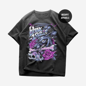 May include: A black short-sleeve t-shirt featuring a graphic print of a snarling wolf, a skull, and purple roses. The text reads Blink 182 and With Vandals and Sharks. A circular logo in the top right corner displays the text MOSHPIT APPARELS.