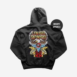 Pode incluir: Um moletom com capuz preto apresentando um gráfico nas costas com o texto Lynyrd Skynyrd e Southern Rock-Roll. O design inclui uma águia-de-cabeça-branca com asas abertas, um escudo circular com uma estrela e fitas amarelas em fundo preto.