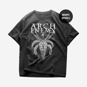 May include: A black short-sleeve t-shirt featuring the white text ARCH ENEMY above a detailed graphic of a multi-horned, multi-eyed goat creature with a skull. A circular black badge in the top right corner displays the white text MOSHPIT APPARELS.