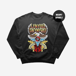 Pode incluir: Uma camisola preta de gola redonda com um gráfico de uma águia-de-cabeça-branca com asas abertas, um escudo circular e faixas amarelas com o texto Lynyrd Skynyrd e Southern Rock-Roll. Um círculo preto com o texto Moshpit Apparels está no canto.