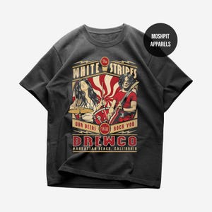 May include: A charcoal grey short-sleeve t-shirt featuring a graphic print of The White Stripes with the text "The White Stripes," "Our Beers Will Roch You," "Brewco," and "Manhattan Beach, California." A black circle with "Moshpit Apparels" is visible.