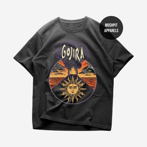 Puede incluir: Una camiseta gris carbón de manga corta con un gráfico circular de una erupción volcánica y un sol estilizado en primer plano. El texto GOJIRA aparece sobre el volcán y un logotipo circular de MOSHPIT APPARELS está en la parte superior derecha.