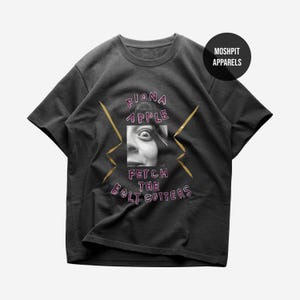 Könnte beinhalten: Ein anthrazitfarbenes Kurzarm-T-Shirt mit einem schwarz-weißen Porträt eines Gesichts, das in einem Quadrat zentriert ist. Der Text FIONA APPLE FETCH THE BOLT CUTTERS ist in Pink gedruckt, und goldene Blitze flankieren das Porträt. MOSHPIT APPARELS steht in einem Kreis.
