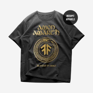 May include: A charcoal gray short-sleeve t-shirt featuring a gold graphic print. The design includes the text AMON AMARTH, a circular knotwork serpent emblem with a central rune-like symbol, and the text THE PURSUIT OF VIKINGS at the bottom.