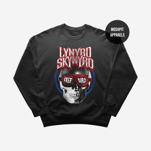Pode incluir: Uma camisola preta de gola redonda com um gráfico de um crânio a usar óculos de aviador vermelhos com o texto "FREE BIRD" nas lentes. Acima do crânio, o texto "LYNYRD SKYNYRD" está impresso em branco e vermelho. Um logótipo circular diz "MOSH PIT APPARELS".