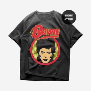 May include: A black short-sleeve t-shirt featuring a graphic print of David Bowie. The design includes the word Bowie in red letters above a circular yellow pop-art style portrait with a red border. A black circle with MOSHPIT APPARELS is visible.