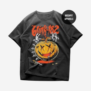 May include: A black short-sleeve t-shirt featuring a graphic of a carved pumpkin with X-shaped eyes and a wide grin. Above the pumpkin is the text "blink-182" in orange. Two white ghost figures, a knife, and a black circle with "MOSH PIT APPARELS" are visible.