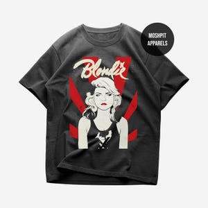 May include: A charcoal gray short-sleeve t-shirt featuring a graphic print of a person with blonde hair and red lips. The text "Blondie" appears in white script above the figure, with red sunburst rays behind. A black circle contains "MOSH PIT APPARELS".