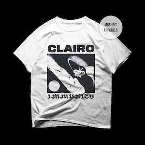 May include: A white short-sleeve t-shirt featuring a black graphic print. The text CLAIRO appears above a square illustration of a moon, stars, and a small creature. The word IMMUNITY is printed below the graphic. A circular MOSHPIT APPARELS logo is visible.
