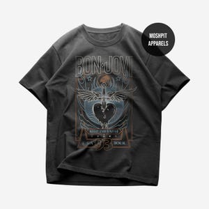 以下が含まれることがあります： BON JOVI、KEEP THE FAITH、USA 93 TOURの文字がプリントされたチャコールグレーの半袖Tシャツ。デザインには、白、青、オレンジの輪郭で描かれた翼のある心臓、短剣、炎のモチーフが含まれています。