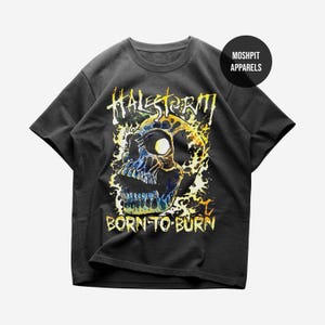 Puede incluir: Una camiseta negra de manga corta con un gráfico de una calavera rodeada de llamas amarillas y azules. El texto "HALESTORM" aparece sobre la calavera y "BORN TO BURN" está impreso debajo. Un logotipo circular dice "MOSH PIT APPARELS".