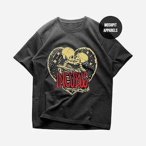 Op de afbeelding: Een antracietgrijs T-shirt met korte mouwen en een afbeelding van twee skeletten die elkaar omhelzen in een hartvorm, omringd door sterren. Het woord INCUBUS staat in dikke rode letters in het midden. Een rond logo toont MOSHPIT APPARELS.