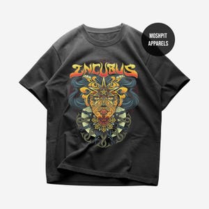 Op de afbeelding: Een antracietgrijs T-shirt met korte mouwen en een grafische print van een gestileerd gezicht met ingewikkelde gele en oranje patronen, donker haar en het woord INCUBUS in gele en rode letters. Een zwarte cirkel bevat de tekst MOSHPIT APPARELS.