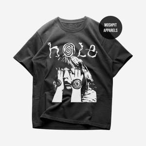 May include: A charcoal grey short-sleeve t-shirt featuring a white graphic print of a person with hands covering their face, displaying spiral designs on the palms and forehead. The word "hole" is printed above, and "MOSH PIT APPARELS" is in a circle.