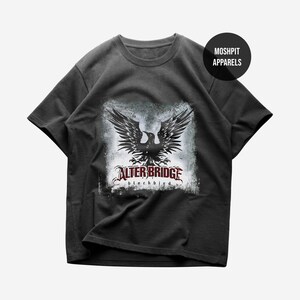 May include: A charcoal gray short-sleeve t-shirt featuring a graphic of a black bird with spread wings over a distressed white square background. The text reads "ALTER BRIDGE" in red and "blackbird" in black. A black circle says "MOSH PIT APPARELS".