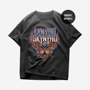 Pode incluir: Uma t-shirt cinzento-carvão de manga curta com um gráfico de uma águia-de-cabeça-branca com asas abertas, duas guitarras elétricas cruzadas e o texto Lynyrd Skynyrd em azul claro. Um logótipo circular preto apresenta o texto MOSHPIT APPARELS.