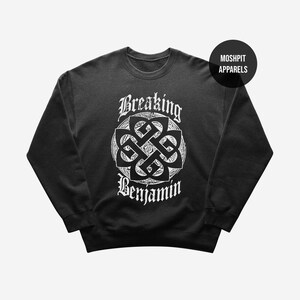 Può includere: Una felpa nera a maniche lunghe con una stampa grafica bianca sul petto. Il design include un emblema circolare a intreccio con il testo Breaking Benjamin sopra e sotto. È visibile un piccolo cerchio nero con il testo MOSHPIT APPARELS.