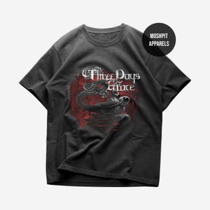 May include: A charcoal gray short-sleeve t-shirt featuring a graphic print of a coiled snake over a distressed red circular background. The text "Three Days Grace" is printed in white above the snake, and a black circle reads "MOSH PIT APPARELS".