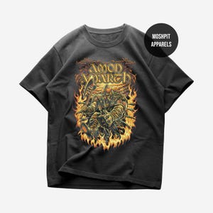 May include: A black short-sleeve t-shirt featuring a graphic print of a Viking warrior holding an axe amidst flames. The text AMON AMARTH is printed above the figure in a stylized font. A circular logo with the text MOSHPIT APPARELS is visible.