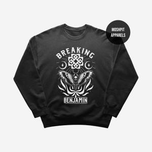Può includere: Una felpa nera a maniche lunghe con una stampa grafica bianca raffigurante una falena con un teschio sul corpo, un nodo celtico, lune crescenti e il testo BREAKING BENJAMIN. Un badge circolare nero nell'angolo riporta la scritta MOSHPIT APPARELS.