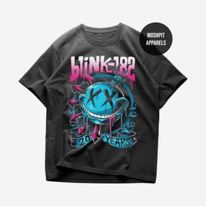 May include: A black short-sleeve t-shirt featuring a graphic print of a blue smiley face with black X eyes and a wide grin. The design includes the text Blink-182, 20 Years, and Moshpit Apparels, with pink and blue arrows and a small rabbit figure.