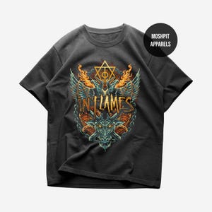 May include: A charcoal gray t-shirt featuring a graphic print of a horned creature, chains, and flames. The text "IN FLAMES" is centered in a stylized font, with a geometric symbol above it. A circular logo reads "MOSH PIT APPARELS" in the corner.