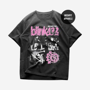 May include: A charcoal gray short-sleeve t-shirt featuring a pink and white graphic print. The design includes the text "blink-182" and "ONE MORE TIME..." along with four panels showing band members, a skeleton, and a circular logo with arrows.
