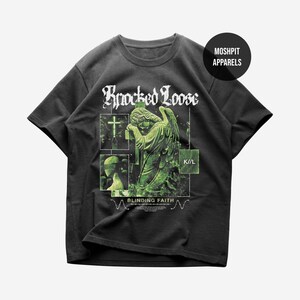 May include: A charcoal gray short-sleeve t-shirt featuring a green graphic print of a weeping angel statue, a cross, and the text Knocked Loose, K//L, and Blinding Faith. A circular black logo in the top right corner reads MOSHPIT APPARELS.