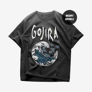 Puede incluir: Una camiseta gris carbón de manga corta con el texto blanco GOJIRA sobre un gráfico circular de una ola rompiendo con detalles en azul y blanco y siluetas de pájaros volando. Se observa un círculo negro con el texto MOSHPIT APPARELS.