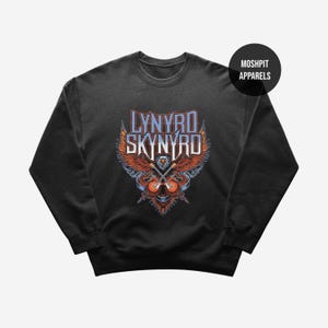 Pode incluir: Uma camisola preta de gola redonda com uma impressão gráfica de uma águia-de-cabeça-branca com asas abertas, duas guitarras elétricas cruzadas e o texto Lynyrd Skynyrd em letras azuis e brancas. Um logótipo circular preto diz Moshpit Apparels.