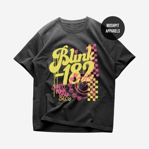 May include: A charcoal gray short-sleeve t-shirt featuring a yellow and pink graphic print. The design includes the text Blink-182, Crappy Punk Rock Since 92, a smiley face icon, and a checkered pattern. A circular logo reads MOSHPIT APPARELS.