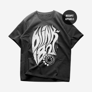 May include: A black short-sleeve t-shirt featuring a large white graphic print of the text Blink-182 in a stylized font with a dripping effect. Below the text is a circular smiley face logo with an X for one eye. The text MOSHPIT APPARELS is visible.
