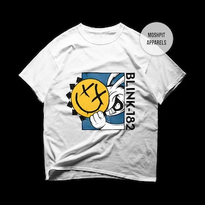May include: A white short-sleeve t-shirt featuring a graphic print of a cartoon rabbit holding a yellow smiley face with X-shaped eyes. The design includes a blue square background, black jagged shapes, and the text BLINK-182 printed vertically in black.