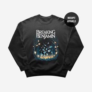 Può includere: Una felpa nera a maniche lunghe con il testo Breaking Benjamin in lettere bianche sopra una grafica di alberi blu, luci scintillanti e frammenti di vetro fluttuanti. Un badge nero circolare riporta la scritta Moshpit Apparels.