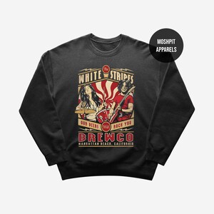 May include: A black crewneck sweatshirt featuring a graphic print of a drummer and a guitarist. The text reads The White Stripes, Our Beers Will Rock You, Brewco, and Manhattan Beach, California. A circular logo in the corner says Moshpit Apparels.