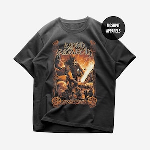 May include: A charcoal gray short-sleeve t-shirt featuring a graphic print of a warrior with a flaming sword and the text AMON AMARTH. A circular black logo in the top right corner displays the white text MOSHPIT APPARELS.