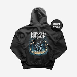 Può includere: Una felpa con cappuccio nera con la scritta Breaking Benjamin in lettere bianche sopra un grafico di frammenti di vetro infranto circondati da alberi blu scuro e sfere gialle luminose. Un logo nero circolare riporta Moshpit Apparels.