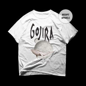 Op de afbeelding: Een wit t-shirt met korte mouwen en een zwarte afbeelding van een walvis die voor een grote maan zwemt. De tekst GOJIRA staat in het zwart boven de walvis, met daaronder FROM MARS TO SIRIUS. Een rond MOSHPIT APPARELS logo is zichtbaar.