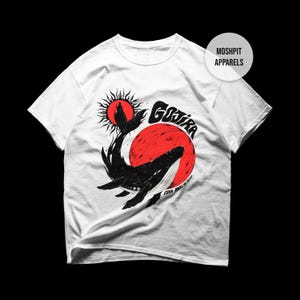 Puede incluir: Una camiseta blanca de manga corta con un gráfico de una ballena negra saltando frente a un gran sol rojo. El diseño incluye el texto "GOJIRA" y "FROM MARS TO SIRIUS" en negro, junto con un icono de sol más pequeño. Un logotipo circular dice "MOSH PIT APPARELS".